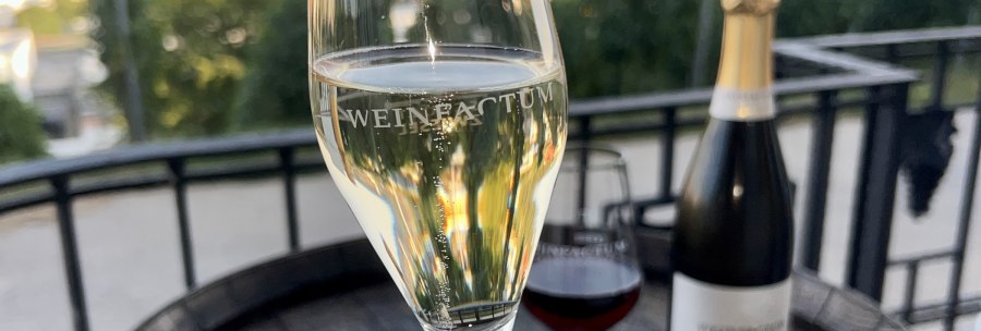 A champagne glass labeled 'Weinfactum' stands on an outdoor table, with a bottle and a wine glass next to it., &copy; Weinfactum Bad Cannstatt
