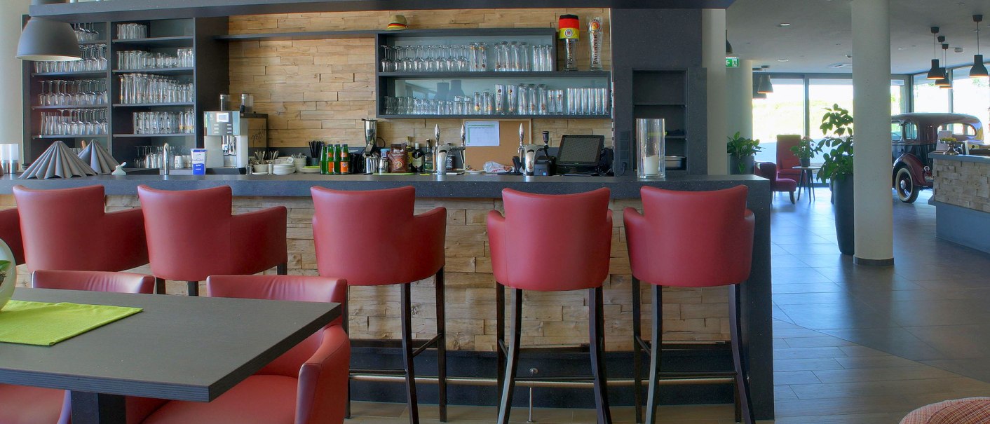 Modern hotel bar with red bar stools, glasses on the shelf and a classic car in the background. Bright, inviting atmosphere., © TOMAS