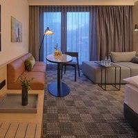 Modern hotel room with sofa, table, bed and large windows. Subtle lighting and stylish decoration create a cozy atmosphere., © Adina Apartment Hotel Stuttgart Modern hotel room with sofa, table, bed and large windows. Subtle lighting and stylish decoration create a cozy atmosphere., © Adina Apartment Hotel Stuttgart