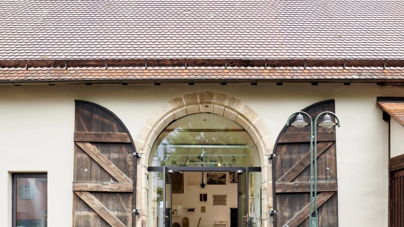 Eingang des Heimatmuseums Plieningen mit großen, offenen Holztüren und einer Glasfront. Innen sind Ausstellungsstücke sichtbar., © StadtPalais / svenweber_repro_ret-min Eingang des Heimatmuseums Plieningen mit großen, offenen Holztüren und einer Glasfront. Innen sind Ausstellungsstücke sichtbar., © StadtPalais / svenweber_repro_ret-min