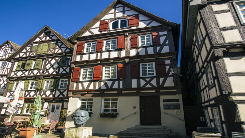 Half-timbered houses in Schorndorf in sunny weather. A bust stands in front of one of the houses. Clear blue sky in the background., © SMG Stuttgart Marketing GmbH - Sarah Schmid Half-timbered houses in Schorndorf in sunny weather. A bust stands in front of one of the houses. Clear blue sky in the background., © SMG Stuttgart Marketing GmbH - Sarah Schmid