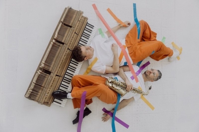 Zwei Männer in orangefarbener Kleidung liegen auf einem weißen Boden, umgeben von bunten Klebebändern. Einer hält ein Saxophon, der andere liegt neben einer Klaviatur., © Renitenztheater Stuttgart e.V. Zwei Männer in orangefarbener Kleidung liegen auf einem weißen Boden, umgeben von bunten Klebebändern. Einer hält ein Saxophon, der andere liegt neben einer Klaviatur., © Renitenztheater Stuttgart e.V.