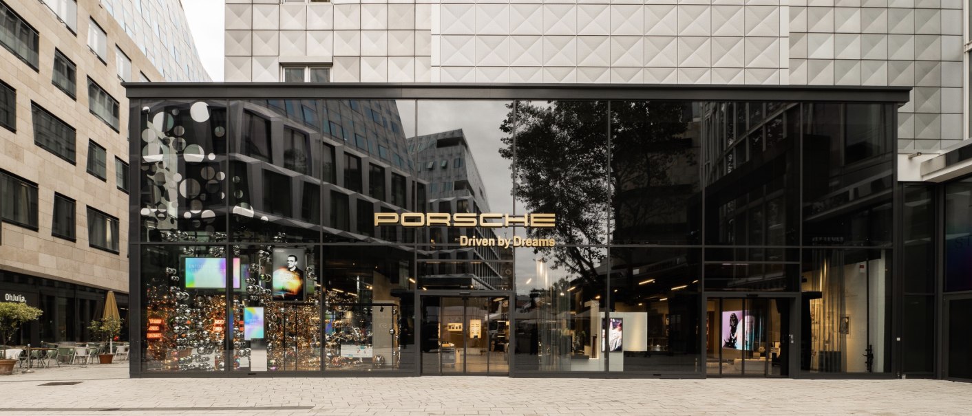 Porsche Brand Store mit gro&szlig;er Glasfassade, umgeben von modernen Geb&auml;uden. Der Schriftzug "Porsche Driven by Dreams" ist prominent sichtbar., &copy; Porsche AG