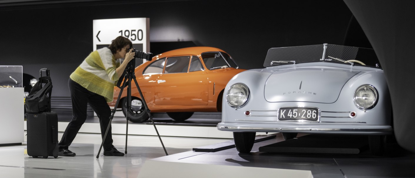 Eine Person fotografiert ein blaues Porsche-Auto in einem Museum. Im Hintergrund steht ein orangefarbenes Auto. Ein Koffer steht neben der Person., © Porsche AG Eine Person fotografiert ein blaues Porsche-Auto in einem Museum. Im Hintergrund steht ein orangefarbenes Auto. Ein Koffer steht neben der Person., © Porsche AG