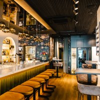 Elegante Bar mit langer Theke, gelben Barhockern und stilvollen Lampen. Die Wände sind mit floralen Mustern dekoriert, Flaschenregale im Hintergrund., © SMG, Sarah Schmid Elegante Bar mit langer Theke, gelben Barhockern und stilvollen Lampen. Die Wände sind mit floralen Mustern dekoriert, Flaschenregale im Hintergrund., © SMG, Sarah Schmid