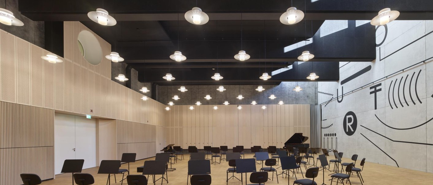 A modern hall with chairs and music stands, light-colored wood paneling, geometric wall patterns, and hanging lamps., &copy; TOMAS