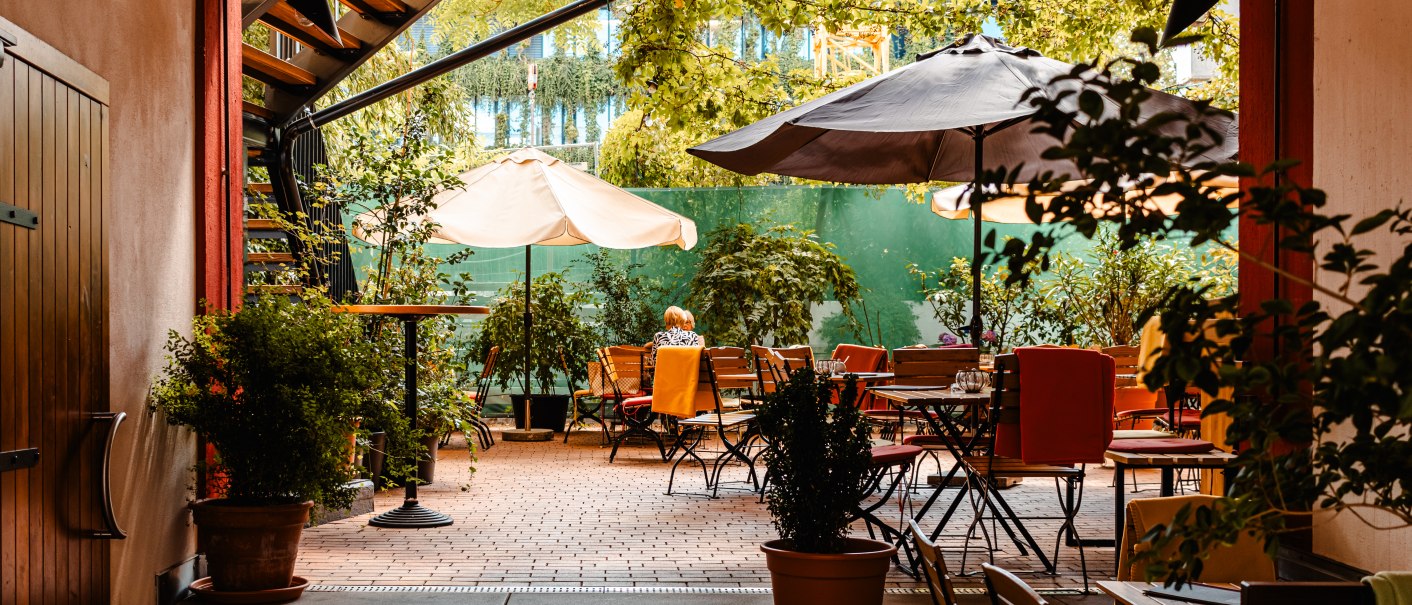 Ein gemütlicher Innenhof eines Cafés mit Tischen, Stühlen und Sonnenschirmen, umgeben von grünen Pflanzen und Bäumen., © SMG_Sarah Schmid