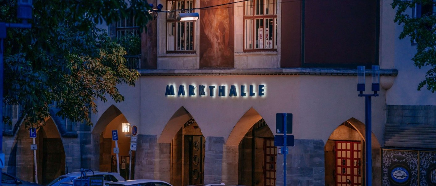 Eingang zur Markthalle bei Nacht, beleuchtetes Schild und Bögen, umgeben von Bäumen und Straßenschildern., © Thomas Niedermüller