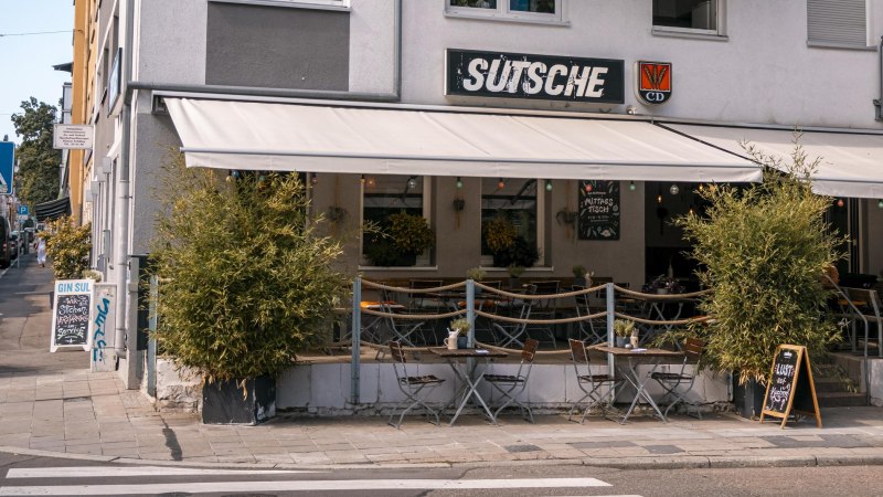 Café 'Sutsche' mit Außenbereich, Tischen und Stühlen auf dem Gehweg, umgeben von Pflanzen., © SMG, Sarah Schmid
