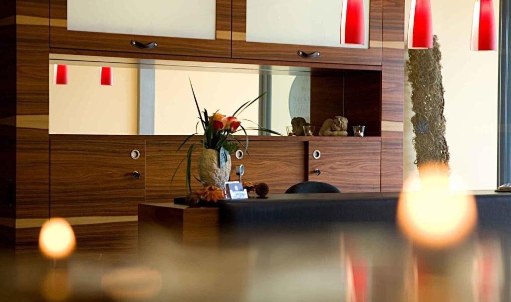 Modern reception desk with wooden paneling, red hanging lamps and floral decorations. Mirrors and decorative objects are visible in the background., © Nödinger Gastro GmbH
