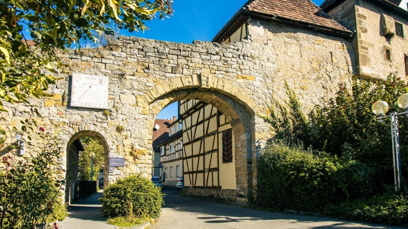 An old town gate in Marbach am Neckar, flanked by half-timbered houses and surrounded by green vegetation under a blue sky., © Stuttgart-Marketing GmbH, Sarah Schmid An old town gate in Marbach am Neckar, flanked by half-timbered houses and surrounded by green vegetation under a blue sky., © Stuttgart-Marketing GmbH, Sarah Schmid