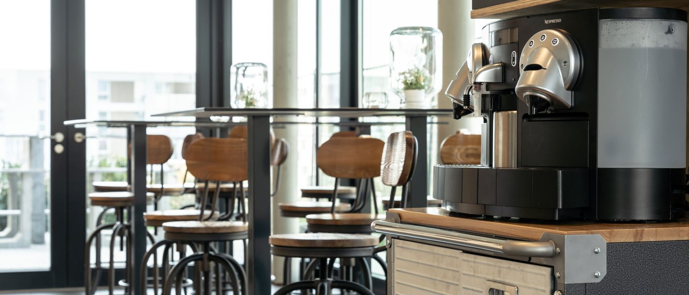 A modern breakfast area with a coffee machine in the foreground and high tables with chairs in the background., © a2 HOTELS A modern breakfast area with a coffee machine in the foreground and high tables with chairs in the background., © a2 HOTELS