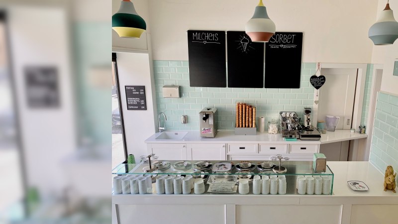 Ice cream parlor with counter, ice cream machine, coffee maker and ice cream cones. Pastel-colored tiles and lamps. Black boards with ice cream offers., © Zur Schleckerei