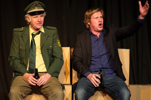 Two men are sitting on cardboard boxes, one wearing a police uniform, the other casual clothes. Both are holding bottles of beer, the man on the right is gesticulating animatedly., © Theaterhaus Stuttgart e.V. Two men are sitting on cardboard boxes, one wearing a police uniform, the other casual clothes. Both are holding bottles of beer, the man on the right is gesticulating animatedly., © Theaterhaus Stuttgart e.V.