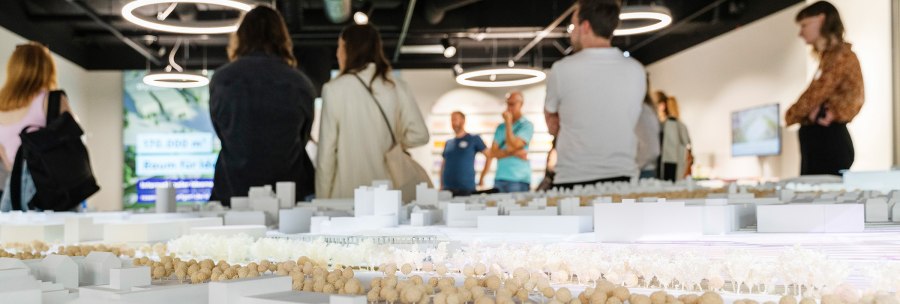 Besucher betrachten ein Architekturmodell in einer Ausstellung. Moderne Deckenleuchten und ein Bildschirm im Hintergrund., &copy; Dominique Brewing