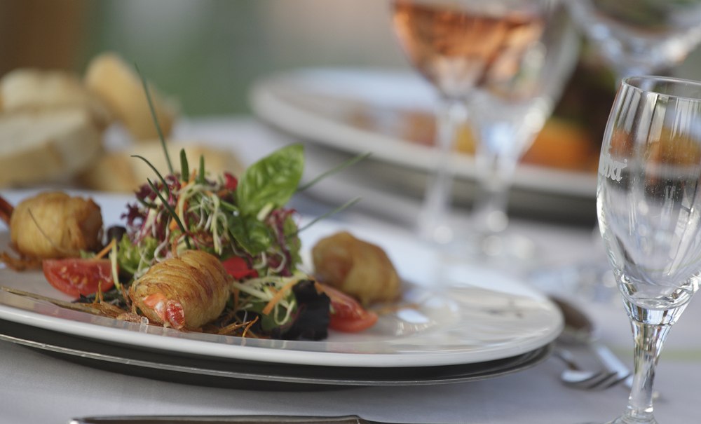 Ein elegantes Gericht mit Garnelen und Salat auf einem weißen Teller, umgeben von Weingläsern auf einem gedeckten Tisch., © Badhotel Restaurant Stauferland