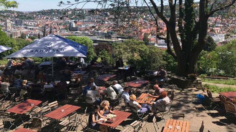 Ein Biergarten auf der Karlshöhe mit Blick auf Stuttgart. Menschen sitzen an Tischen unter Sonnenschirmen, umgeben von Bäumen und einer Stadtansicht., © Stuttgart-Marketing GmbH