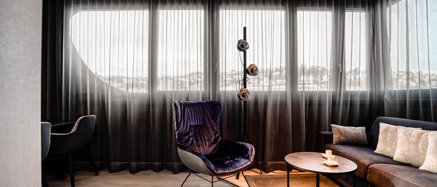 Modernes Wohnzimmer mit großen Fenstern, einem blauen Sessel, Sofa, Couchtisch und Stehlampe. Helle Vorhänge lassen Licht herein., © CLOUD N°7 APARTMENTS GmbH