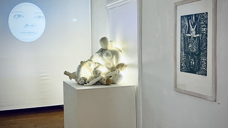 Eine Kunstinstallation zeigt eine Gesichtprojektion, beleuchtete Puppen auf einem Podest und ein gerahmtes Bild an der Wand., © Min Bark Eine Kunstinstallation zeigt eine Gesichtprojektion, beleuchtete Puppen auf einem Podest und ein gerahmtes Bild an der Wand., © Min Bark