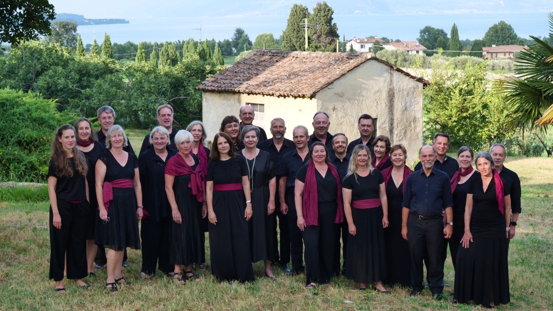 Gruppe von Menschen in schwarzer Kleidung mit roten Akzenten vor einem kleinen Gebäude in einer grünen Landschaft., © KCBW