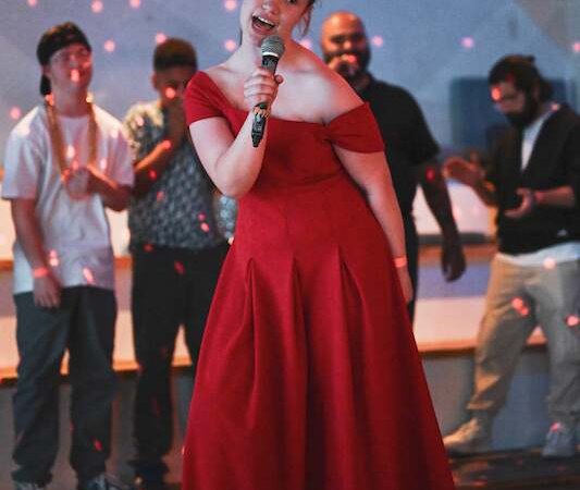 Eine Frau in einem roten Kleid singt auf einer B&uuml;hne. Im Hintergrund stehen mehrere Personen. Die Atmosph&auml;re ist festlich mit bunten Lichtern., &copy; Rosenau Kultur e.V.