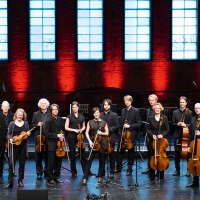 Das Stuttgarter Kammerorchester posiert auf einer Bühne vor einer roten Backsteinwand mit großen Fenstern. Die Musiker halten ihre Instrumente., © Reiner Pfisterer Das Stuttgarter Kammerorchester posiert auf einer Bühne vor einer roten Backsteinwand mit großen Fenstern. Die Musiker halten ihre Instrumente., © Reiner Pfisterer