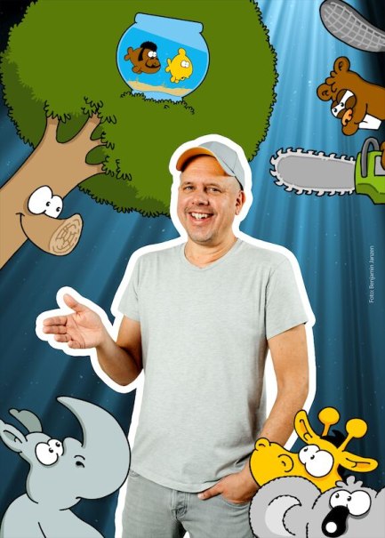 Ein Mann in einem grauen T-Shirt steht lächelnd vor einer Collage aus Cartoon-Tieren und -Objekten, darunter ein Nashorn, ein Baum und ein Fischglas., © Theaterhaus Stuttgart e.V. Ein Mann in einem grauen T-Shirt steht lächelnd vor einer Collage aus Cartoon-Tieren und -Objekten, darunter ein Nashorn, ein Baum und ein Fischglas., © Theaterhaus Stuttgart e.V.