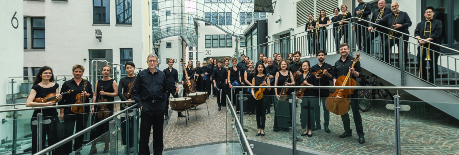 Die Gaechinger Cantorey posiert in einem modernen Innenhof. Die Musiker tragen schwarze Kleidung und halten verschiedene Instrumente., &copy; Martin F&ouml;rster