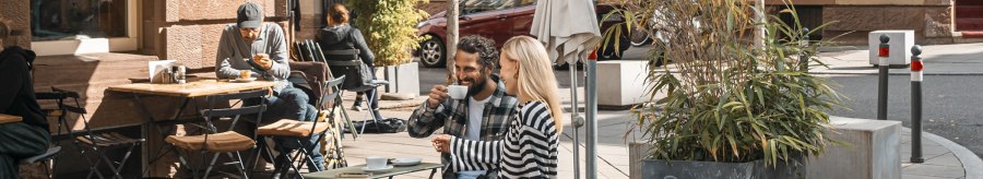 People enjoying coffee in front of a caf&eacute; in the sun. A couple is sitting on a bench, others at tables. Plants and parasols decorate the scene., &copy; SMG, Sarah Schmid