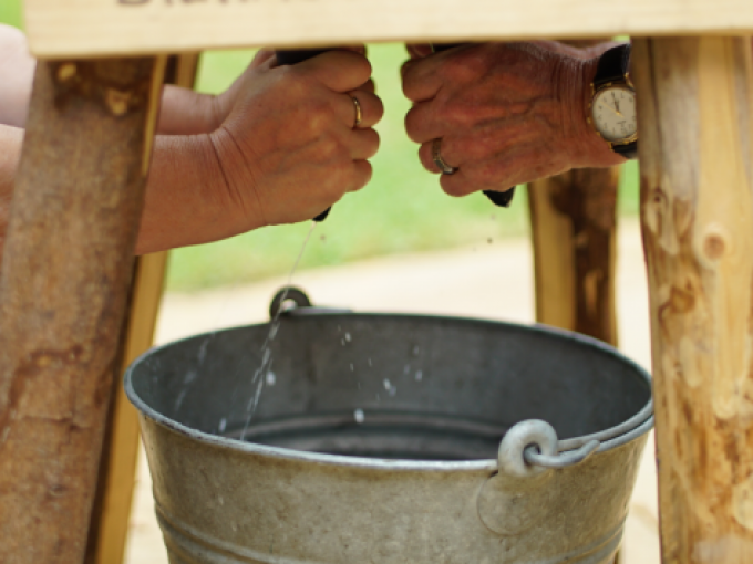 Zwei Personen melken eine Kuhattrappe, Wasser tropft in einen Metalleimer. Beide tragen Ringe, eine Person trägt eine Uhr., © Cool-Tours StattReisen Zwei Personen melken eine Kuhattrappe, Wasser tropft in einen Metalleimer. Beide tragen Ringe, eine Person trägt eine Uhr., © Cool-Tours StattReisen
