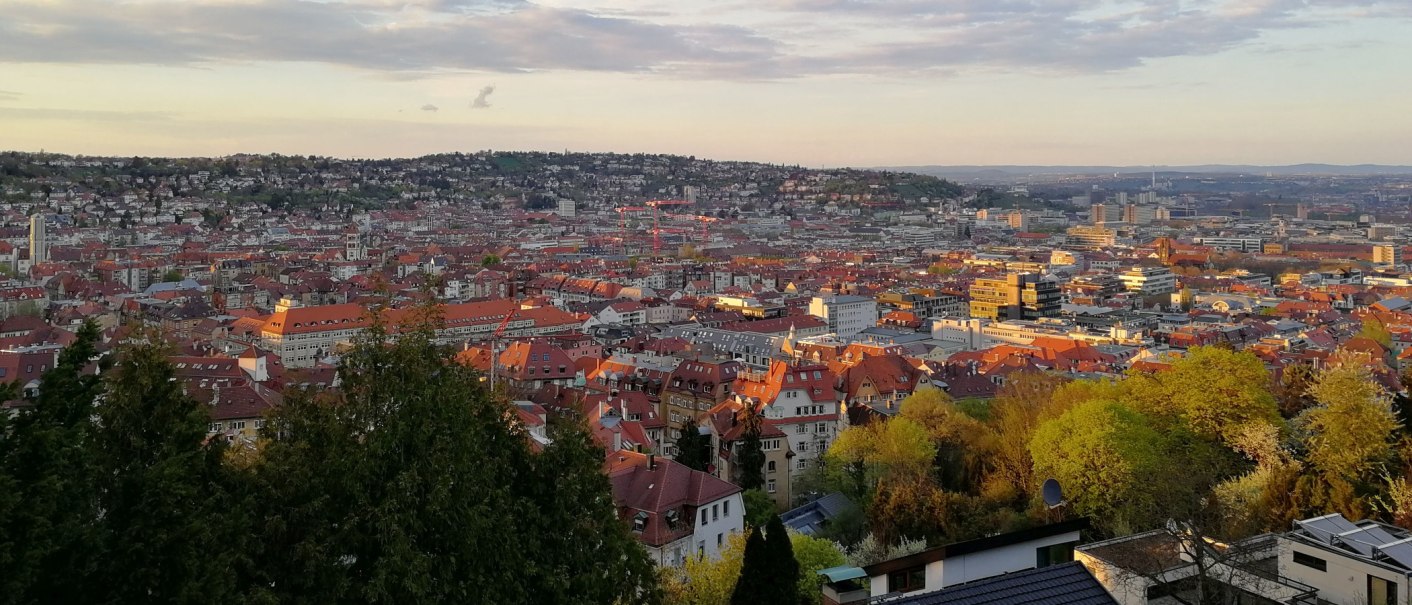 Panoramic view of Stuttgart at sunset, with red roofs and green trees in the foreground. The sky is slightly cloudy., © Stuttgart-Marketing GmbH Panoramic view of Stuttgart at sunset, with red roofs and green trees in the foreground. The sky is slightly cloudy., © Stuttgart-Marketing GmbH