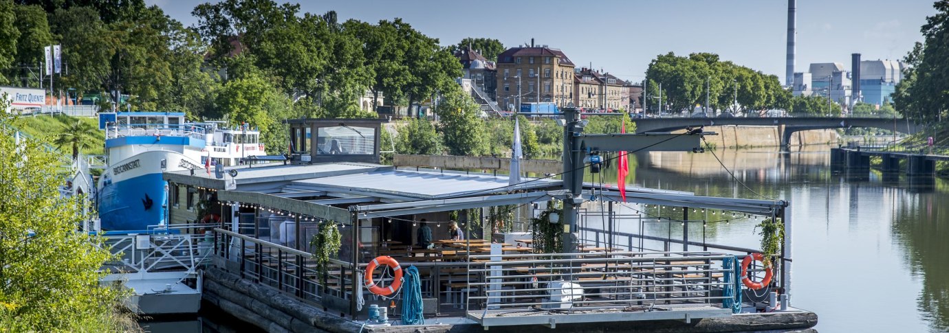 Ein schwimmendes Restaurant auf dem Neckar, umgeben von gr&uuml;ner Vegetation und st&auml;dtischen Geb&auml;uden im Hintergrund., &copy; all copyrights are reserved by maks richter