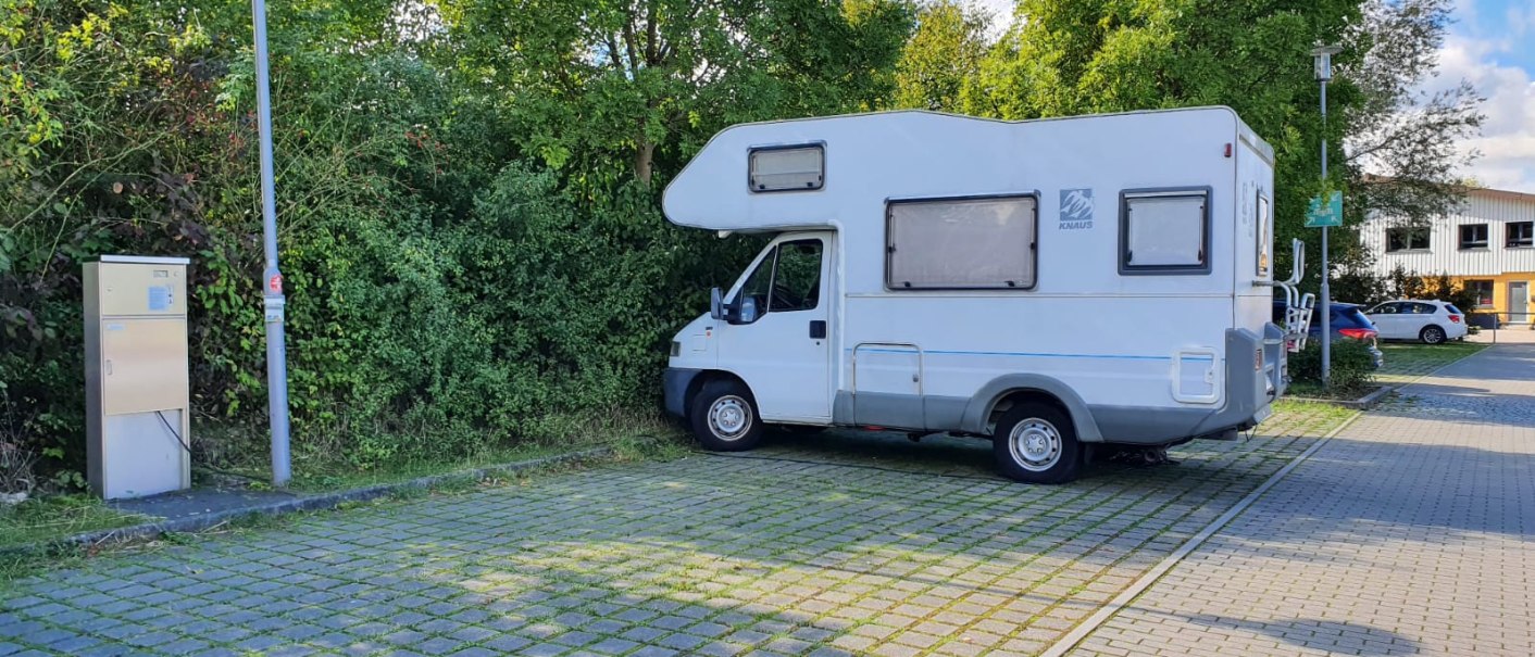 Ein Wohnmobil steht auf einem gepflasterten Stellplatz neben einer Ladestation, umgeben von Bäumen und einem Gebäude im Hintergrund., © Stadt Schorndorf