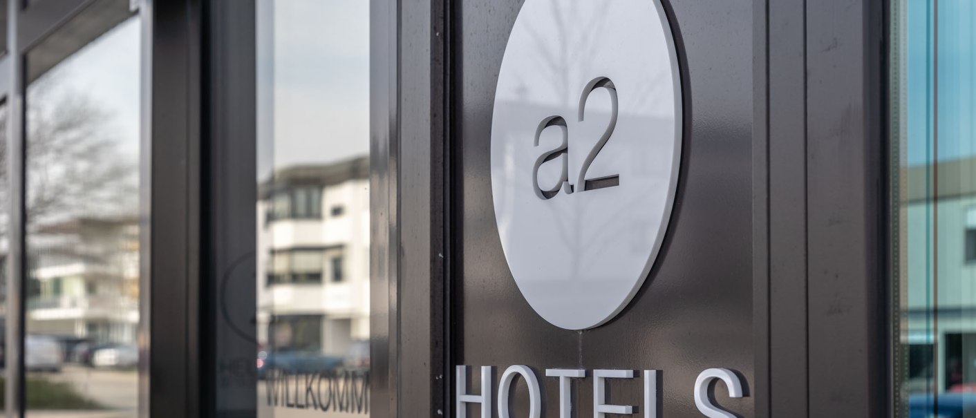 Close-up of the a2 HOTELS entrance with round logo on the glass front. Modern buildings are visible in the background., © a2 HOTELS Close-up of the a2 HOTELS entrance with round logo on the glass front. Modern buildings are visible in the background., © a2 HOTELS