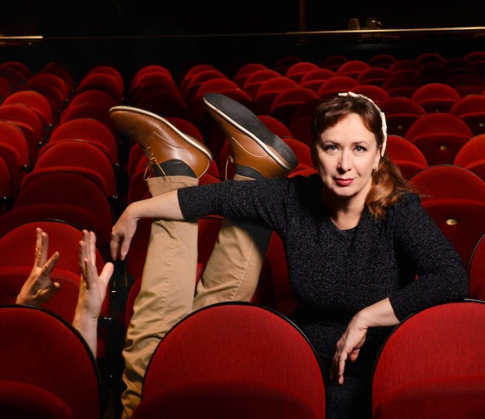 Woman sitting in a theater auditorium while a person's legs and hands protrude from between the red seats next to her., © Rosenau Kultur e.V. Woman sitting in a theater auditorium while a person's legs and hands protrude from between the red seats next to her., © Rosenau Kultur e.V.