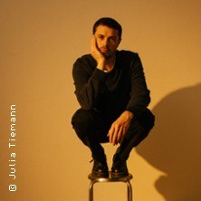 Ein Mann sitzt nachdenklich auf einem Hocker, umgeben von einem warmen, orangefarbenen Licht. Er stützt seinen Kopf auf eine Hand., © links im Bild Ein Mann sitzt nachdenklich auf einem Hocker, umgeben von einem warmen, orangefarbenen Licht. Er stützt seinen Kopf auf eine Hand., © links im Bild