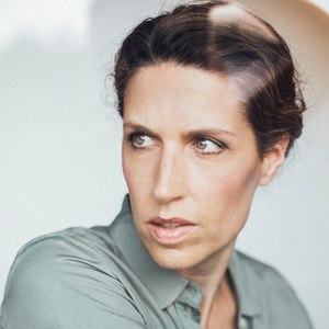 A woman with a serious expression is looking to the side. She is wearing a gray shirt and has dark hair pinned up., © Renitenztheater Stuttgart e.V. A woman with a serious expression is looking to the side. She is wearing a gray shirt and has dark hair pinned up., © Renitenztheater Stuttgart e.V.