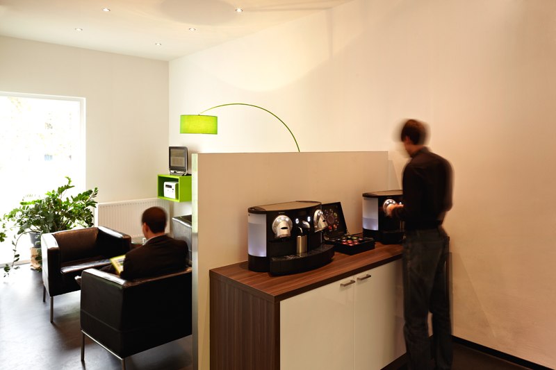 Modern lobby with coffee machine on a cupboard, two people, one sitting, the other standing. Light-colored walls, green lamp and plants., &copy; a2 HOTELS