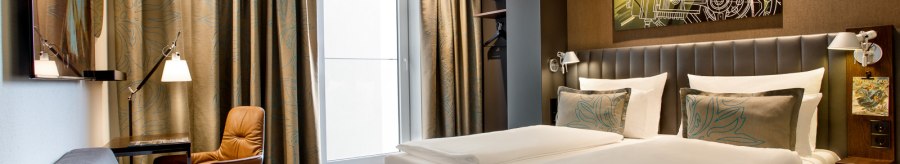 Modernes Hotelzimmer mit Doppelbett, Sofa, Schreibtisch und großem Fenster. An der Wand hängt ein Kunstwerk mit einem Auto-Motiv., © TOMAS