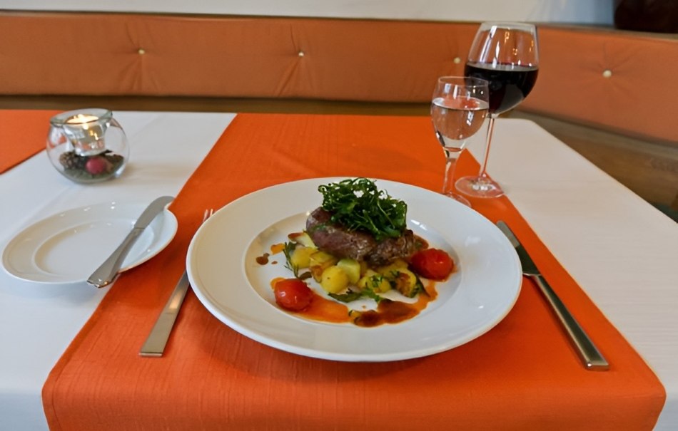 An elegantly laid table with a plate of meat, vegetables and sauce. Next to it is a glass of red wine and water. The tablecloth is orange and white., © Hotel Neuwirtshaus An elegantly laid table with a plate of meat, vegetables and sauce. Next to it is a glass of red wine and water. The tablecloth is orange and white., © Hotel Neuwirtshaus