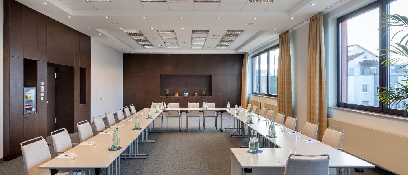 Moderner Konferenzraum mit U-f&ouml;rmiger Tischanordnung, St&uuml;hlen und Wasserflaschen. Gro&szlig;e Fenster lassen viel Licht herein, braune Vorh&auml;nge und Pflanzen dekorieren den Raum., &copy; NH Hotel Stuttgart Airport