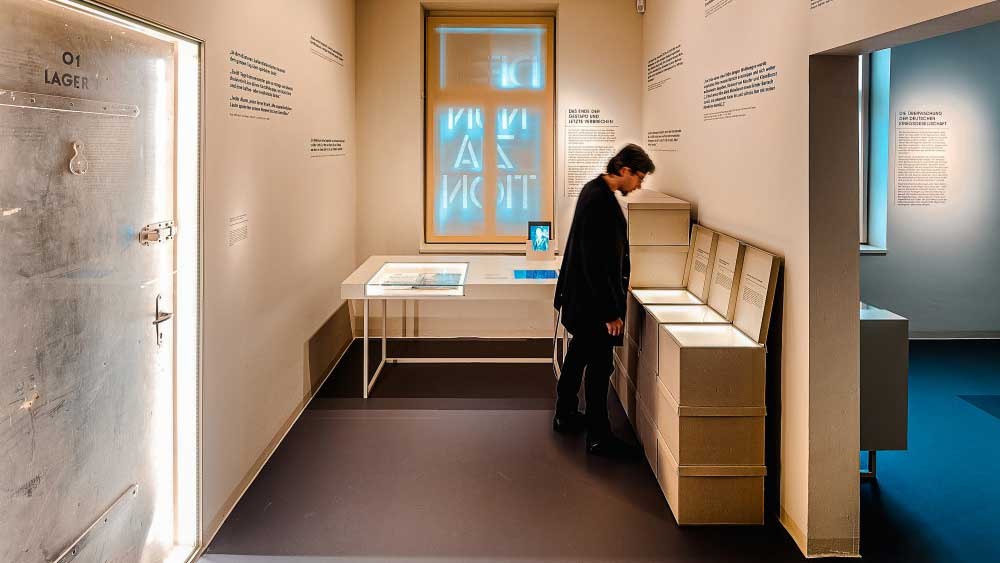 Ein Museumsraum mit einer Person, die Exponate betrachtet. An der Wand sind Texte und ein beleuchtetes Fenster zu sehen. Links eine T&uuml;r mit der Aufschrift '01 Lager'., &copy; Haus der Geschichte Baden-W&uuml;rttemberg