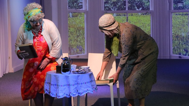 Zwei Frauen in Kostümen stehen auf einer Bühne. Eine hält ein Buch, die andere richtet einen Stuhl. Auf dem Tisch stehen Kaffeekanne und Tassen., © Foto_Katja Ritter Zwei Frauen in Kostümen stehen auf einer Bühne. Eine hält ein Buch, die andere richtet einen Stuhl. Auf dem Tisch stehen Kaffeekanne und Tassen., © Foto_Katja Ritter