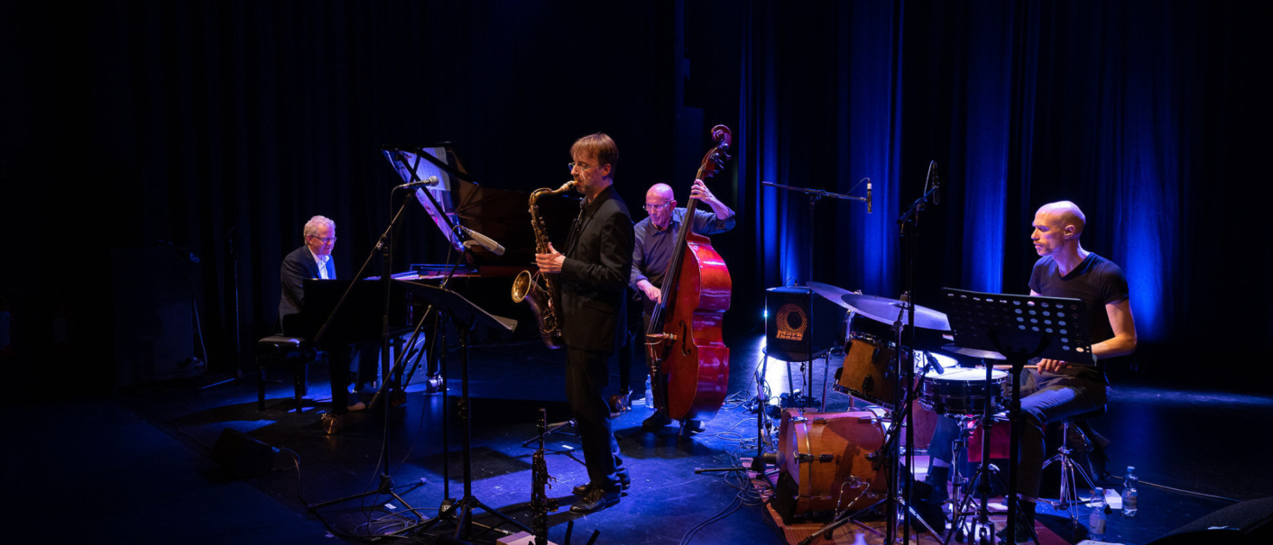 Eine Jazzband spielt auf einer Bühne. Ein Saxophonist, Pianist, Kontrabassist und Schlagzeuger sind in blauem Licht zu sehen., © Bettina Meister