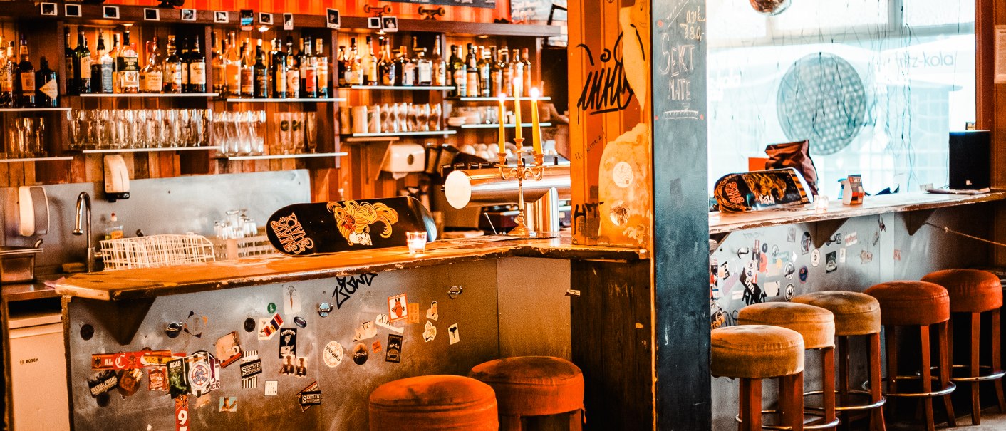 Gemütliche Bar mit Barhockern, einer Theke voller Flaschen und Gläser. Die Wände sind mit Stickern dekoriert, und eine Tafel zeigt Getränkepreise., © Mata Hari