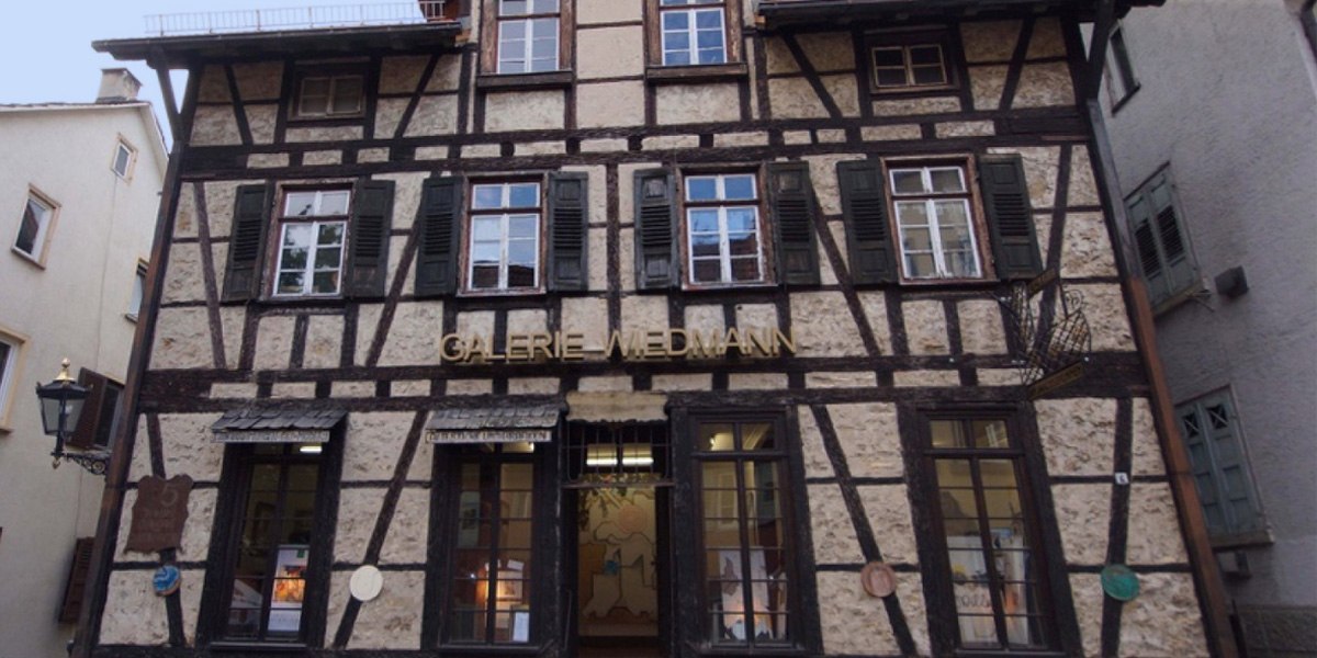 Fachwerkhaus mit der Aufschrift 'Galerie Wiedmann'. Das Gebäude hat mehrere Fenster mit grünen Fensterläden und eine Laterne an der Seite., © Galerie Wiedmann