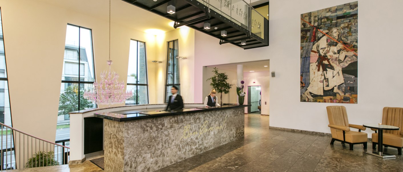 Moderne Hotelrezeption mit Marmortresen, großem Kronleuchter, Kunstwerk an der Wand und zwei Angestellten. Helle, stilvolle Einrichtung., © ARCOTEL Hotels Moderne Hotelrezeption mit Marmortresen, großem Kronleuchter, Kunstwerk an der Wand und zwei Angestellten. Helle, stilvolle Einrichtung., © ARCOTEL Hotels