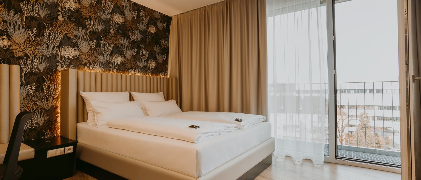 Modernes Hotelzimmer mit Doppelbett, eleganter Tapete und Zugang zu einem Balkon. Helle Vorhänge und Holzboden ergänzen das Design., © TOMAS