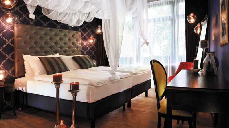 Elegantes Schlafzimmer mit Himmelbett, dunklen Möbeln, Kerzen und stilvoller Dekoration. Große Fenster lassen viel Licht herein., © TOMAS Elegantes Schlafzimmer mit Himmelbett, dunklen Möbeln, Kerzen und stilvoller Dekoration. Große Fenster lassen viel Licht herein., © TOMAS