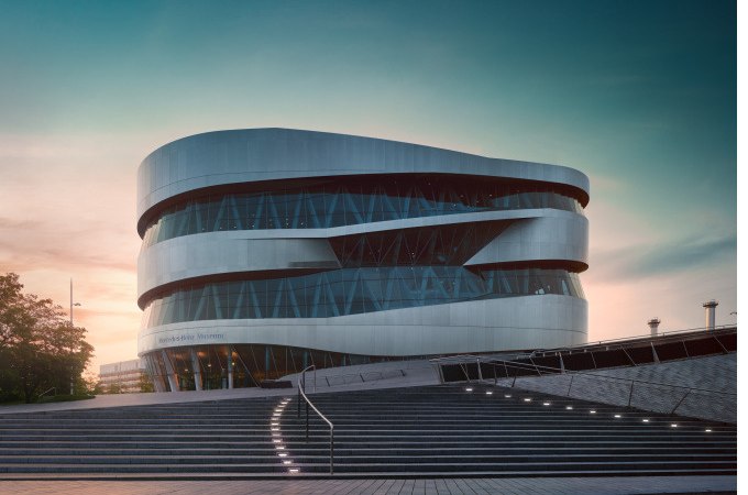 Das Mercedes-Benz Museum mit seiner modernen, geschwungenen Architektur bei Sonnenuntergang, umgeben von Treppen und einem klaren Himmel., © Mercedes-Benz Heritage